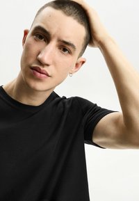 Schwarzes, strukturiertes T-Shirt mit kurzen Ärmeln und rundem Ausschnitt, getragen von einer Person mit kurzen Haaren und einem kleinen Ohrstecker.