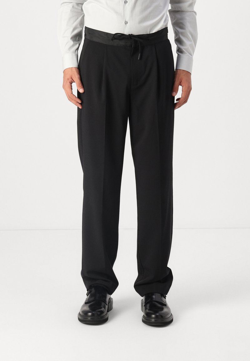 Zwarte, op maat gemaakte broek met een gladde textuur, voorzien van een trekkoord in de taille en plooien, gecombineerd met gepolijste zwarte schoenen.