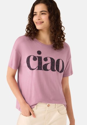 Młoda kobieta z kręconymi włosami, ubrana w malinowy t-shirt z napisem "ciao" i jasnobeżowe spodnie, uśmiechająca się i patrząca w bok.