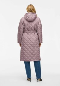 Žena v prešívanom mauve kabáte s kapucňou a pásom, modrých džínsoch a čiernych lodičkách, stojí obrátená chrbtom na bielom pozadí.