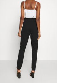 Svarta skräddarsydda byxor med hög midja och smal passform, kombinerat med en vit topp med spaghettistövlar, visad bakifrån.