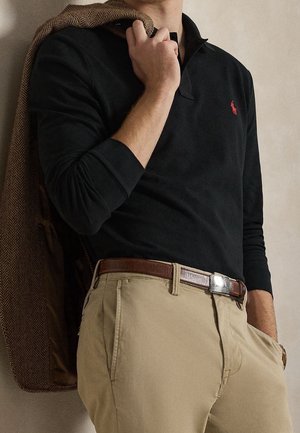 Homme portant une chemise noire à manches longues et un pantalon beige tient une veste marron en chevrons sur l'épaule, montrant une ceinture en cuir marron et un logo rouge.