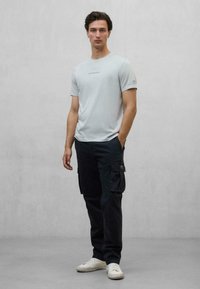 Camiseta de manga corta gris claro con diseño de texto; combinada con pantalones cargo negros con bolsillos laterales; el modelo lleva zapatillas blancas.