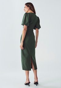 Grünes, tailliertes Midikleid mit Puffärmeln, einem Schlitz im Rücken und glatter Textur. Getragen mit schwarzen, hohen Sandalen, Model blickt von der Kamera weg.