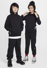 Felpe con cappuccio nere dotate di tasche frontali, abbinate a pantaloni della tuta neri. Entrambi indossano scarpe sportive nere e bianche con dettagli rossi.