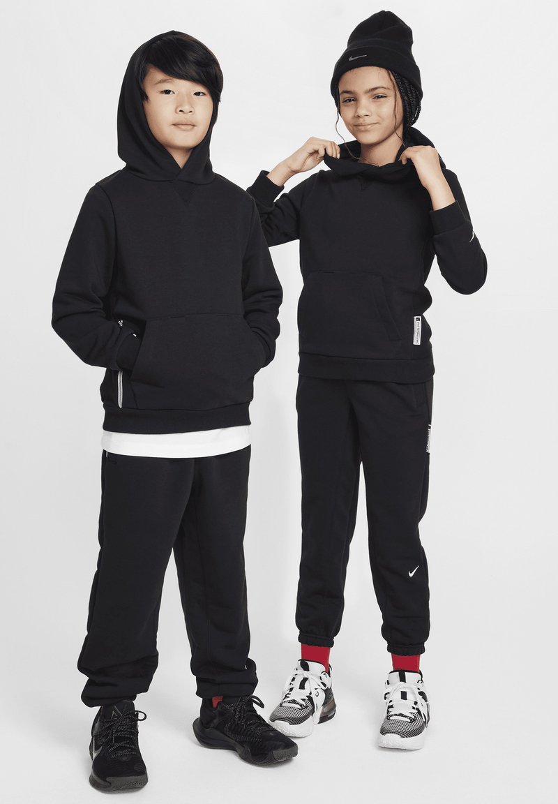 Felpe con cappuccio nere dotate di tasche frontali, abbinate a pantaloni della tuta neri. Entrambi indossano scarpe sportive nere e bianche con dettagli rossi.