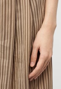 Δεξί χέρι που ακουμπά απαλά σε μπεζ φόρεμα με κάθετες καφέ ρίγες, σε απλό ανοιχτό φόντο.