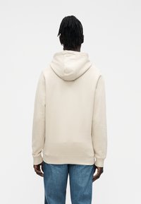 Felpa beige con cappuccio regolabile con coulisse, polsini e orlo a costine. Tessuto liscio e vestibilità comoda, abbinata a pantaloni di denim blu.