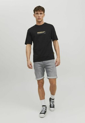 Svart kortärmad t-shirt med guldtext, i kombination med ljusgrå shorts med uppvikta ärmar. Modellen har på sig svarta sneakers och vita strumpor.