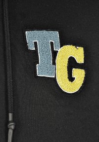 Svart sweatshirt med texturerad applikation av bokstäverna "T" i blått och "G" i gult. Utformad med dragsko och slitstarkt tyg.