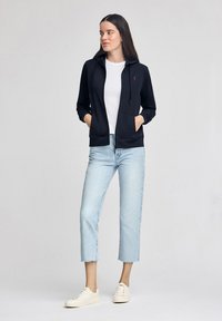 Sudadera negra con cremallera y bolsillos, combinada con una camiseta blanca y vaqueros claros con puños. El calzado son zapatillas deportivas de colores claros.