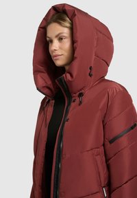 Red puffer jacket with a large hood, zippered front, and black hardware accents. Features a smooth, shiny texture and quilted design.