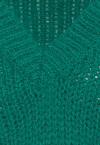 Türkise Strickware mit einem strukturierten Zickzackmuster. Das Material wirkt weich und leicht flauschig, mit einem v-förmigen Ausschnitt.