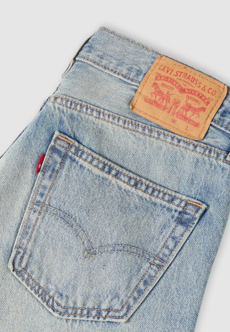 Lyseblå denimjeans med en karakteristisk brun læderlabel ved taljebåndet, der har en rød fane på baglommen og klassiske syninger.