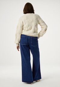 Crème trui met textuur hartpatronen, geribde manchetten, gecombineerd met wijde donkerblauwe jeans. Eenvoudige achtergrond voor focus op de kleding.