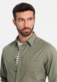 Camicia button-up verde oliva con colletto a punta, taschino sul petto e una texture sottile, indossata sopra una maglietta a righe bianche e blu navy.