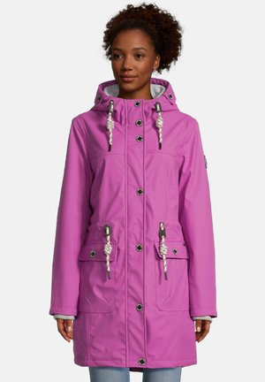 Vrouw die een lange paarse gewatteerde winterjas met capuchon draagt, met witte trekkoorden en steekzakken aan de voorkant, staand tegen een effen achtergrond.