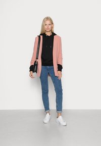 Blazer lungo rosa, felpa con cappuccio nera, jeans skinny blu e sneakers bianche. Il modello tiene una borsa a tracolla e si trova su un pavimento grigio.