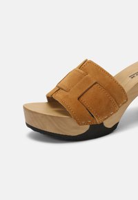 Bruine suède slide sandaal met een brede band, houten blokhak en zwarte rubberen zool. Beschikt over gestikte details op de band.