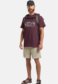 Bordeaux t-shirt met een grafische print, gecombineerd met beige shorts. Draag het met een grijze rugzak en zwarte sandalen. Geplaatst tegen een eenvoudige witte achtergrond.