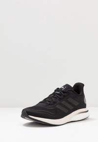 Chaussure de course Adidas noire avec semelle blanche, trois bandes sur le côté et lacets, présentée légèrement en angle sur une surface blanche.