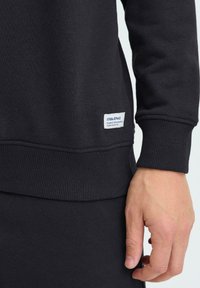 Gros plan d'une personne portant un sweat-shirt noir avec des poignets et un ourlet côtelés, montrant une étiquette rectangulaire blanche près de la taille.