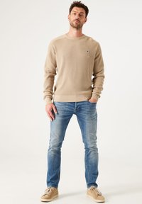 Pull beige côtelé avec un col rond, associé à un jean bleu clair et des baskets en daim beige. Texture remarquable et coupe décontractée.