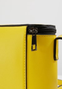 Sac en simili cuir jaune avec une bordure de fermeture éclair noire et une forme rectangulaire. Il est doté d'un tirette de fermeture éclair argentée et d'un design compact.