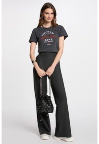 T-shirt graphique gris avec design d'ailes rouges, associé à un pantalon noir taille haute. Les accessoires comprennent un sac seau noir texturé avec des chaînes.