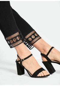 Zwarte broek met een geometrische kantafwerking aan de zoom en een paar zwarte suède sandalen met blokhak en enkelbandjes.