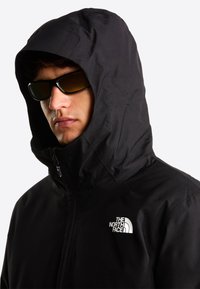 Άνδρας που φοράει μαύρο μπουφάν North Face με κουκούλα σηκωμένη και μαύρα γυαλιά ηλίου, στρεφόμενος ελαφρώς αριστερά μπροστά σε λευκό φόντο.