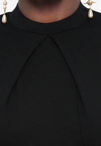 Gros plan d'un vêtement noir à col haut avec un détail de pli triangulaire, porté par une personne à la peau foncée, avec des boucles d'oreilles pendantes en or et perles.