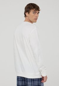 Biała polarowa bluza z długimi rękawami, okrągłym dekoltem i ściągaczem na dole. W komplecie z niebieskimi, w kratę spodniami piżamowymi.