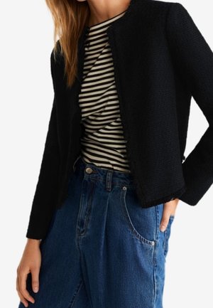Femme portant une veste texturée noire sur une chemise rayée noir et blanc, associée à un jean bleu taille haute, debout avec une main dans la poche.