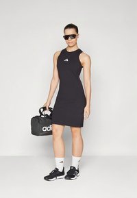 Vestido preto sem mangas feito de tecido elástico, com decote redondo, logo da adidas, combinado com ténis pretos e uma bolsa de ginásio ao mesmo estilo.