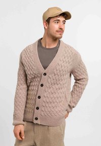 Cardigan beige lavorato a maglia con motivo a treccia, dotato di scollatura a V, cinque bottoni neri, polsini a costine e una vestibilità comoda sopra una camicia grigia.