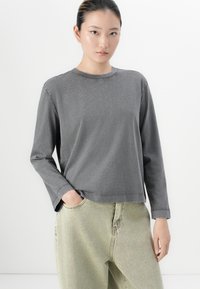 Woman with dark hair in a low bun wearing a faded gray long-sleeve shirt and light green high-waisted jeans, hand in pocket, neutral background.