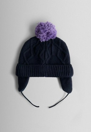 Pletená tmavomodrá čiapka s okrúhlym, huňatým fialovým pom-pomom na vrchu; má uši zakrývajúce chlopne a šnúrky na bezpečné upevnenie; textúrovaný vzor s vrkočmi.
