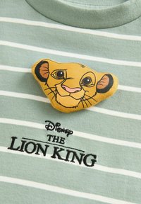 T-shirt a righe verde chiaro con una badge stampata raffigurante il volto di Simba giallo. Sotto, un testo ricamato nero recita "Disney IL RE LEONE".