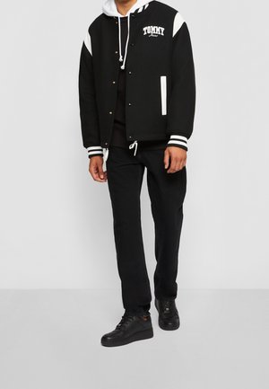 Chaqueta varsity negra con detalles blancos, texto "TOMMY", puños a rayas, combinada con pantalones negros y zapatillas negras. Look deportivo casual.