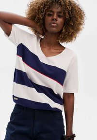 Korte mouwen, V-hals sweater in crème met marineblauwe en rode strepen. Gebreide textuur met geperforeerd detail en een geweven merklabel aan de onderrand.