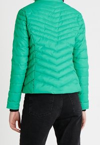 Personne portant une veste matelassée ajustée bleu sarcelle avec des coutures en chevron et un jean noir, vue de dos sur un fond uni.