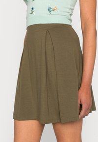 Saia pregueada verde-oliva, feita de um tecido macio. Apresenta um design evasê com uma bainha que cai acima do joelho, exibindo uma textura suave.