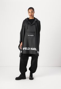 Svart poncho i lätt material med huva. Har en framficka och en vit "KARL LAGERFELD" logga längs fållen. Stylar med svarta byxor och stövlar.