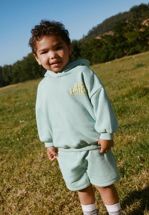 Junges Kind mit lockigem Haar, das einen hellgrünen Kapuzenpullover und Shorts trägt, steht auf Gras mit Bäumen und klarem Himmel im Hintergrund.