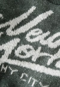 Pull tricot gris avec un motif de texte graphique blanc. Le tissu est texturé et duveteux, avec un col côtelé.