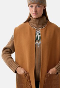 Brauner Wollmix-Rollkragenpullover unter einem beigen ärmellosen Mantel mit glatter Textur; der Mantel verfügt über zwei gesteppte Taschen.
