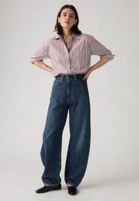 Chemise boutonnée rayée maroon et blanche avec les manches roulées, associée à un jean bleu à taille haute et jambes larges, et des ballerines noires.