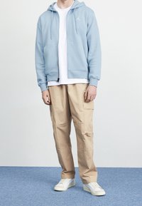 Ljusblå zip-up hoodie med kängurufickor, vit t-shirt under, beige cargobyxor och vita sneakers. Enkel design med minimal märkning.