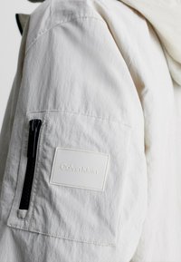 Kreeminvärinen hihapocket, jossa on musta vetoketju ja valkoinen Calvin Klein -logopatch, tekstuurilla varustetussa takissa.